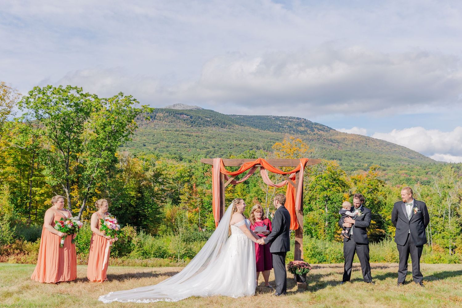 Wedding ceremony under rustic wooden arch with coral draping against mountain landscape.
