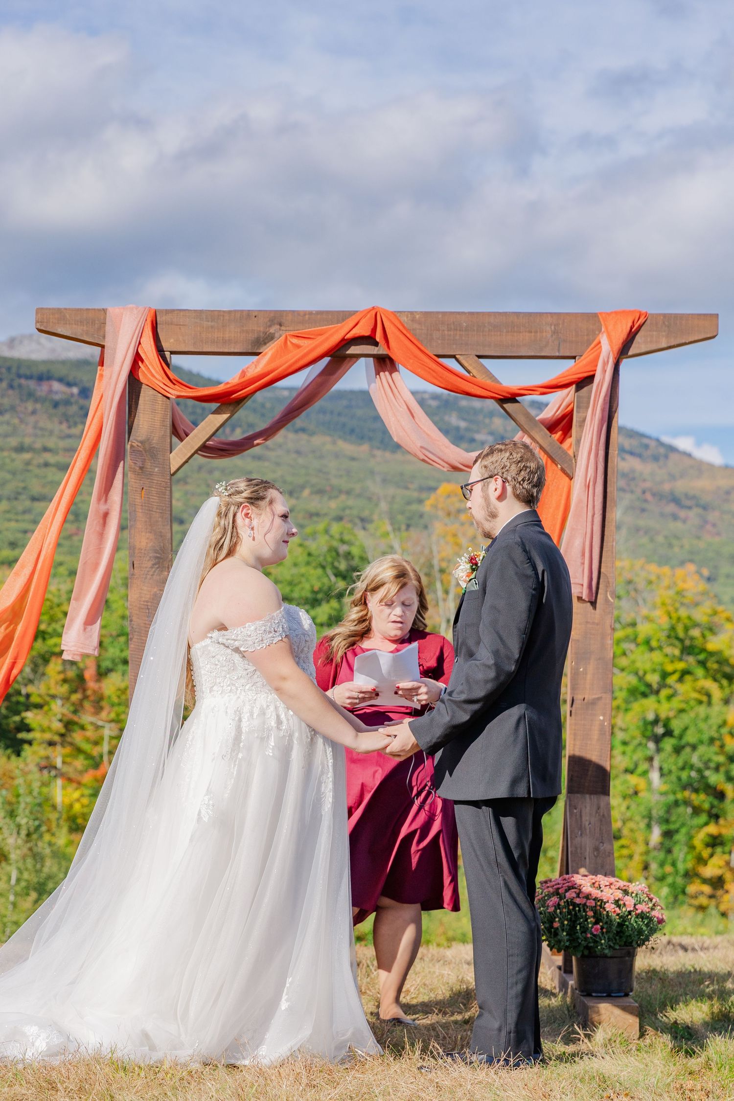 Wedding ceremony under wooden arch with coral draping against autumn mountain backdrop.