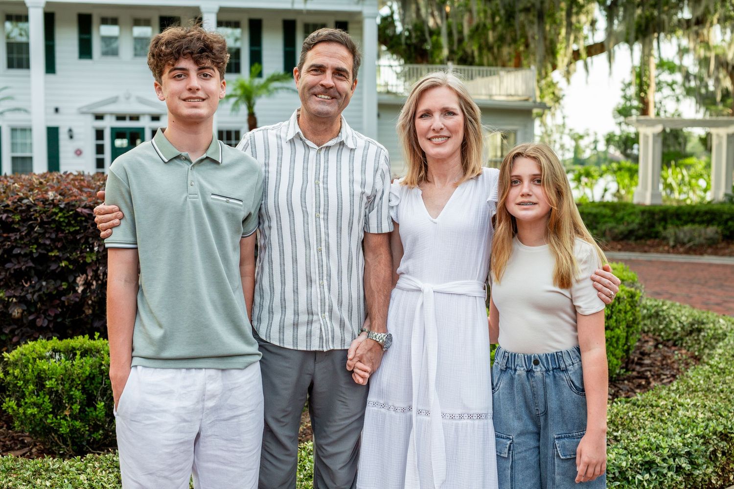 Family members wearing coordinated casual outfits in white, blue and grey stand together in an outdoor park setting.