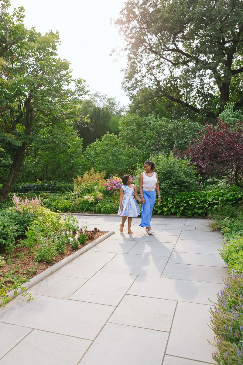 Mom and daughter walking together with sunlit garden backdrop in Central Park