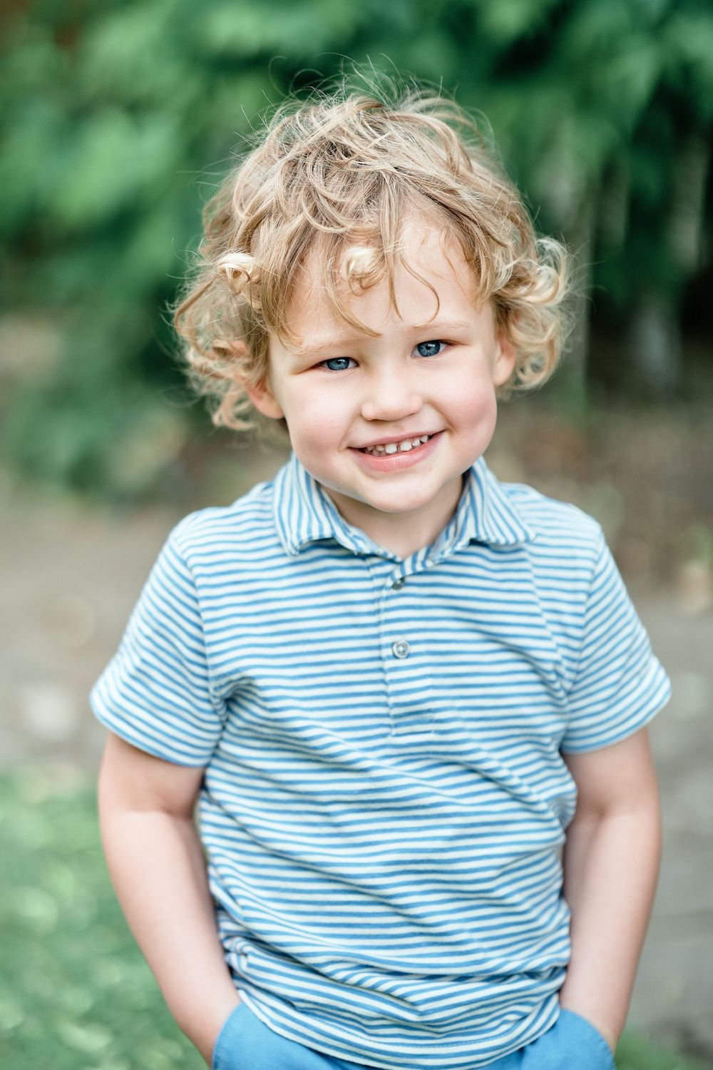 Curly-haired child in blue and white striped polo shirt smiles brightly against blurred green outdoor background.