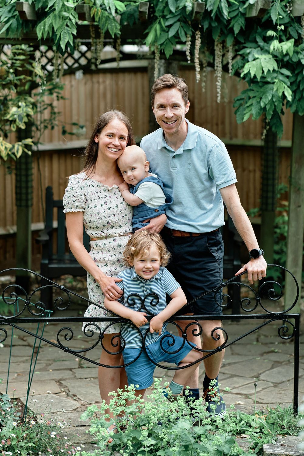Family poses together in a lush garden setting with ornate iron gate and wooden fence backdrop.