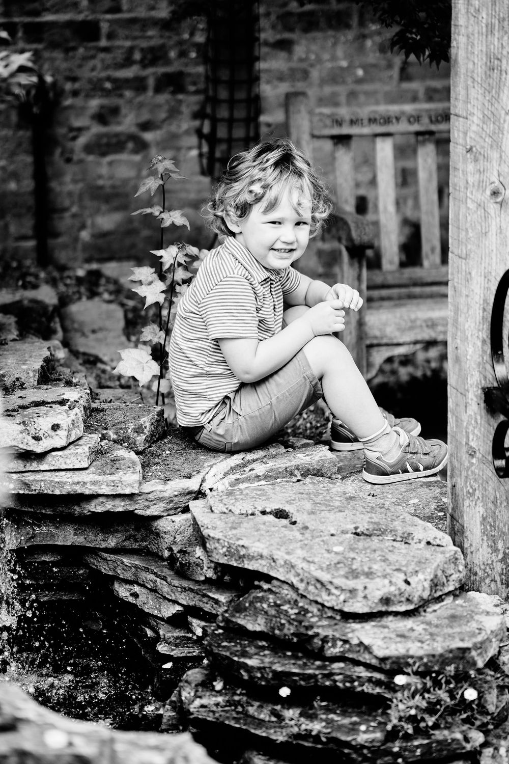 Black and white photo of a child sitting on stone steps in a casual pose wearing a striped shirt and sneakers.