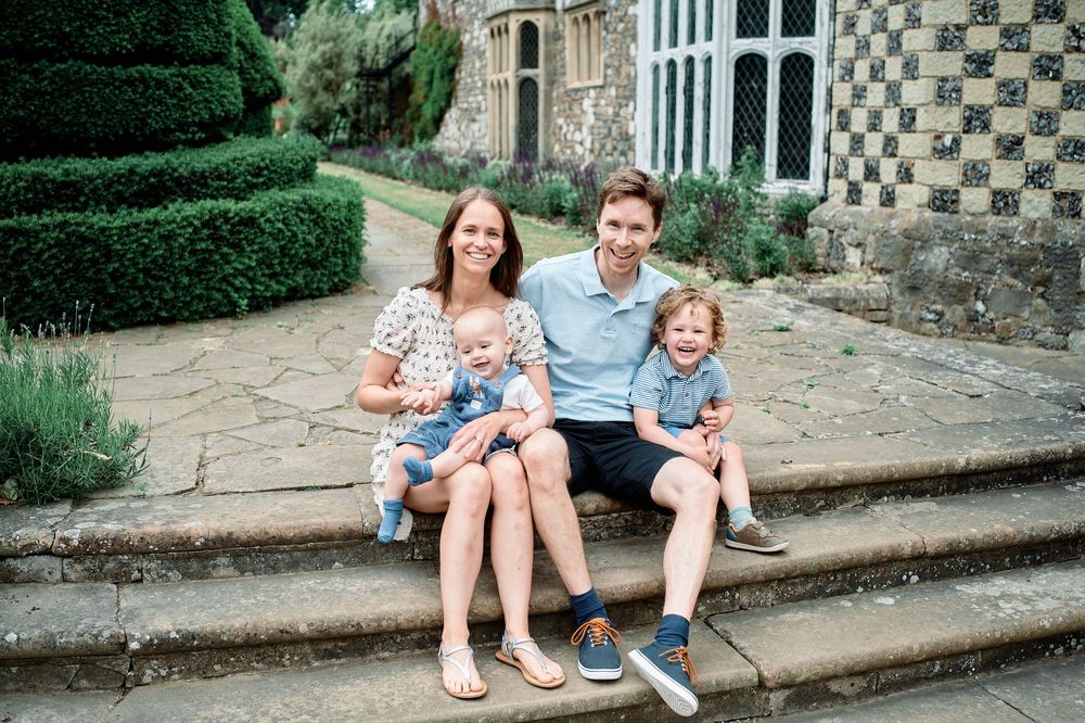 A family of four sits together on stone steps outside a historic building with manicured hedges in the background.
