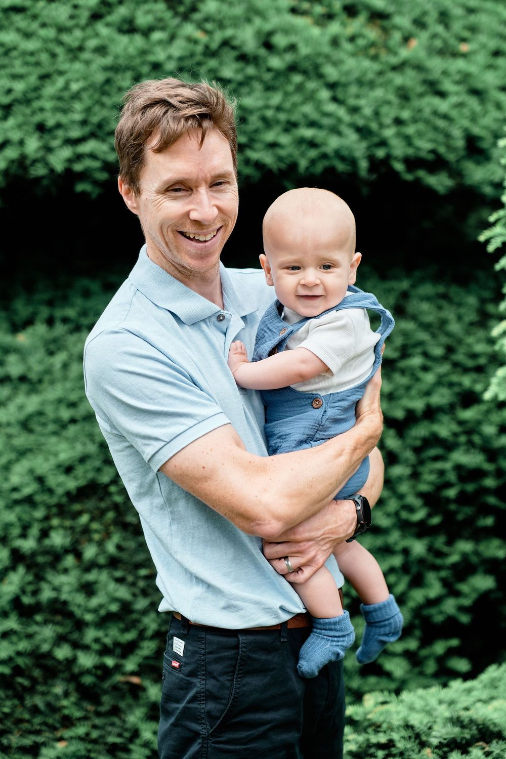 A parent and baby share a joyful moment together outdoors against green foliage background.