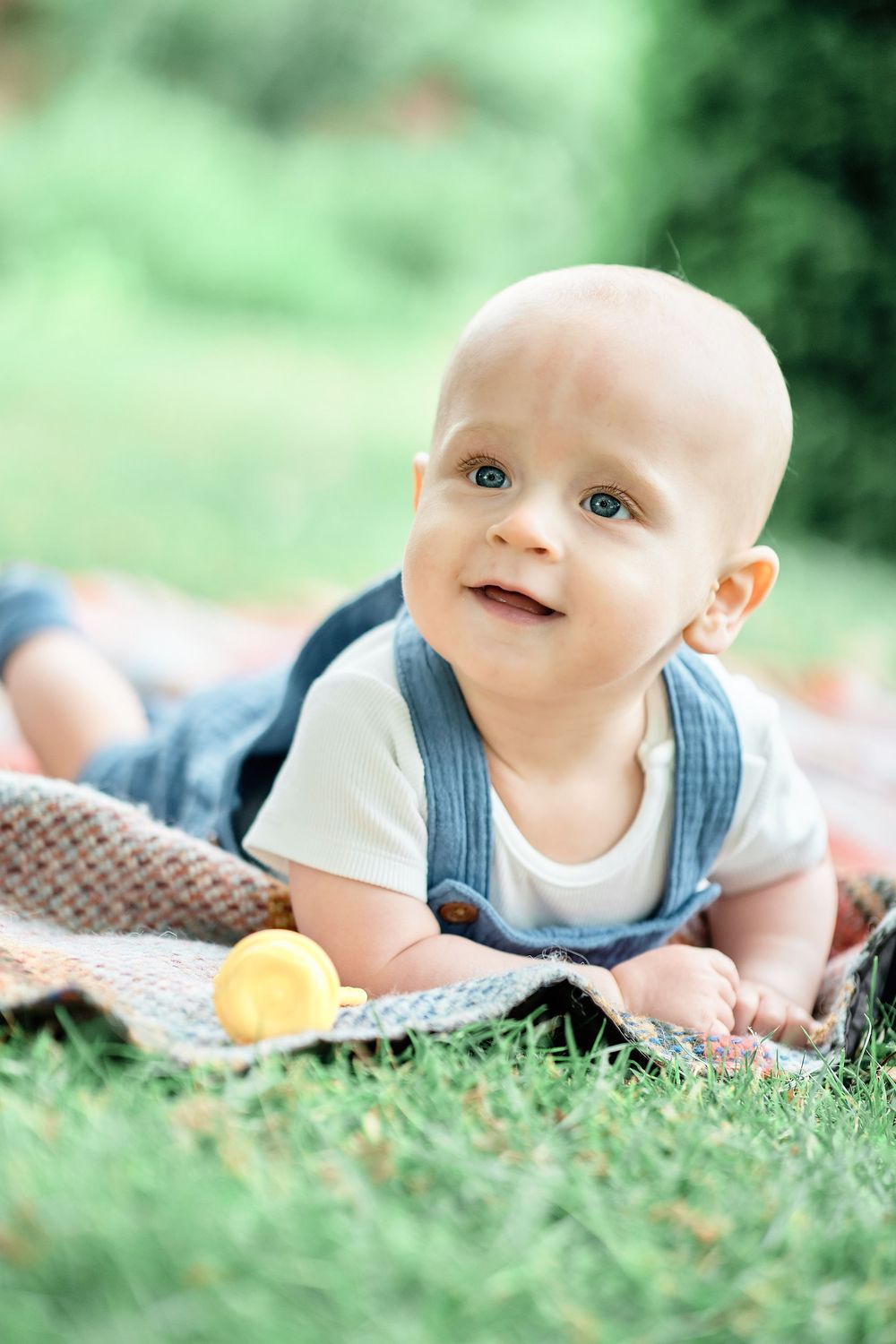 Baby in blue overalls lying on blanket in grass outdoors during summer with a yellow toy.
