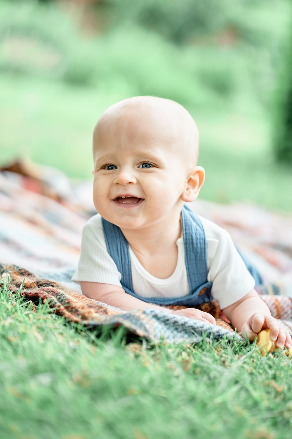 Happy baby in blue overalls lying on a patterned blanket in the grass while smiling during outdoor photoshoot.