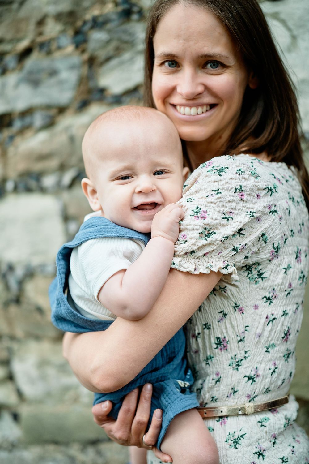 A happy baby wearing blue is held close while both smile brightly against a stone wall background.