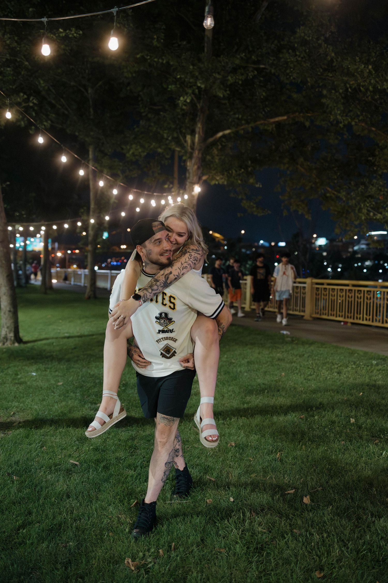 A romantic proposal scene unfolds on a grassy lawn in front of a stone building with someone kneeling and their partner standing.