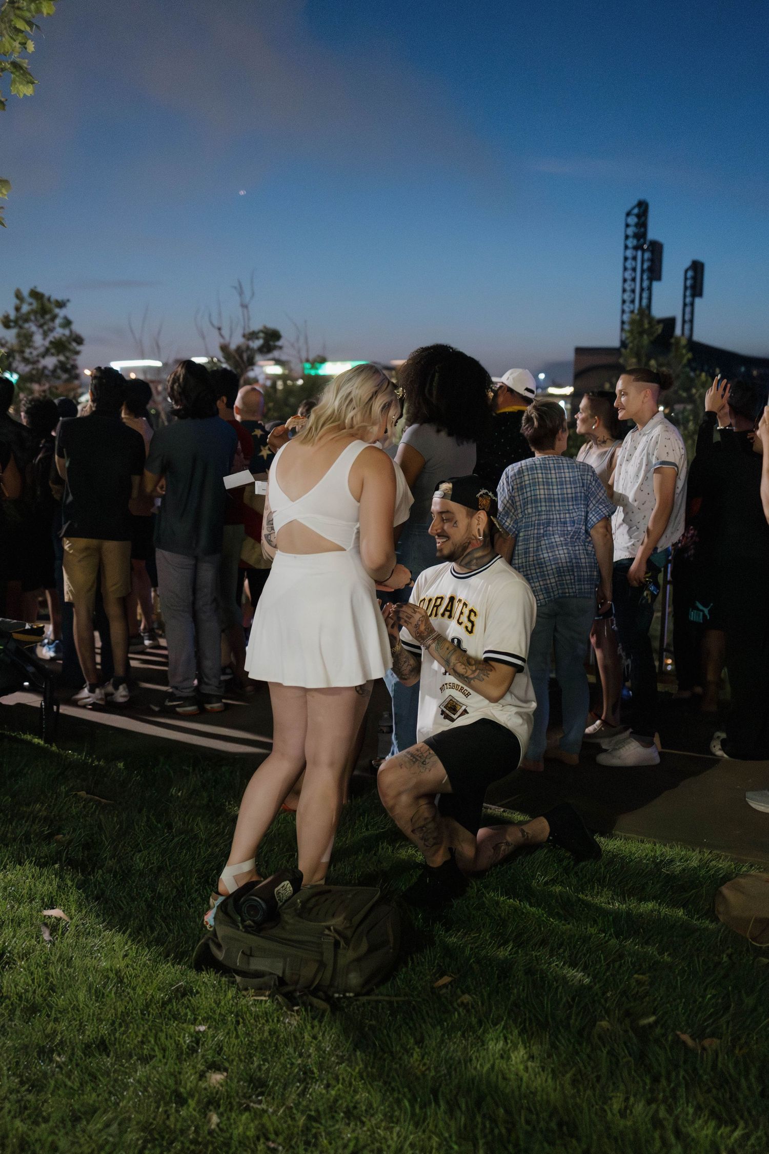 A romantic proposal scene unfolds on a grassy lawn in front of a stone building with someone kneeling and their partner standing.