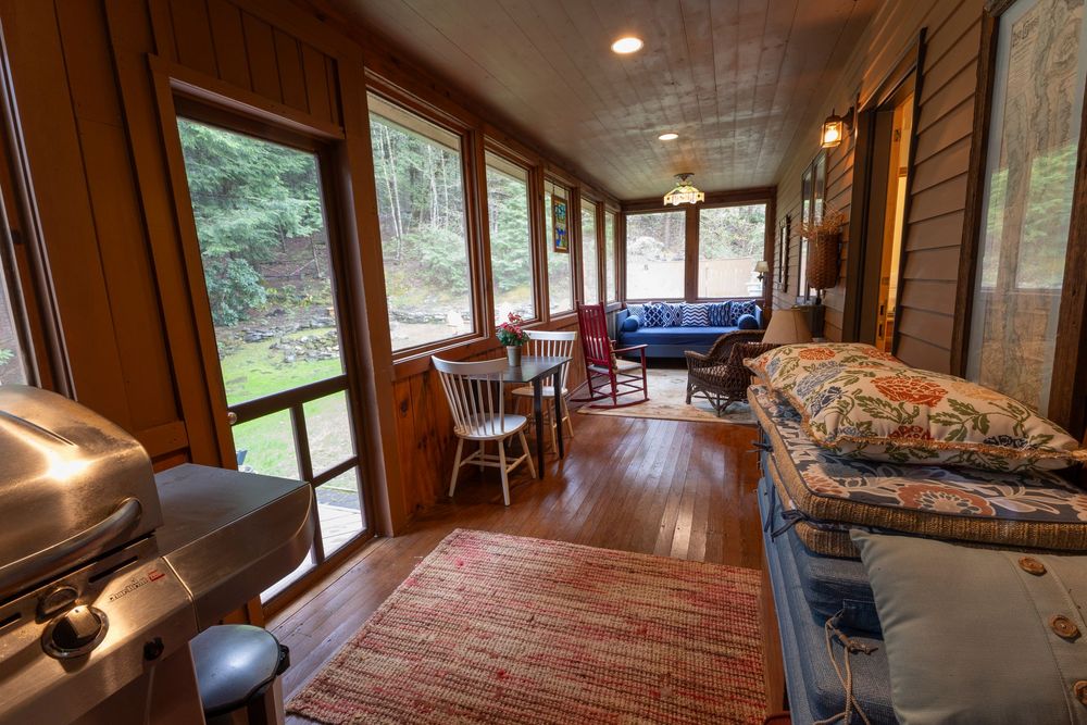 Rustic enclosed porch with wooden floors, screened windows, and comfortable seating overlooking nature.