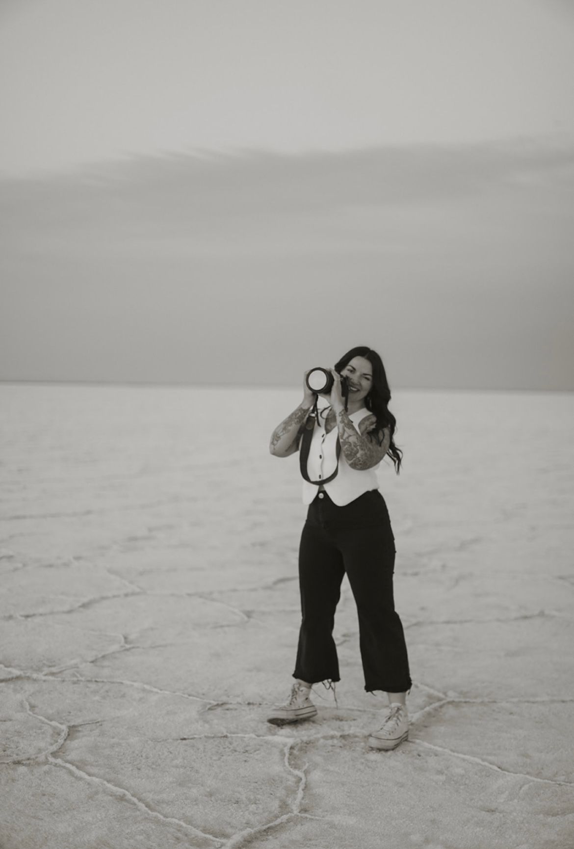 A black and white photo showing Lauren from Terra Firma Photography smiling with her camera on a salt flat landscape.