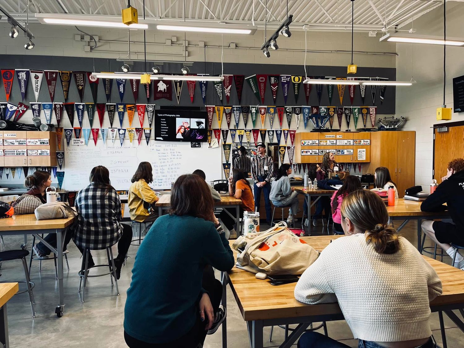 Students work at wooden tables in a modern cafeteria space decorated with colorful pennant banners hanging from the ceiling.