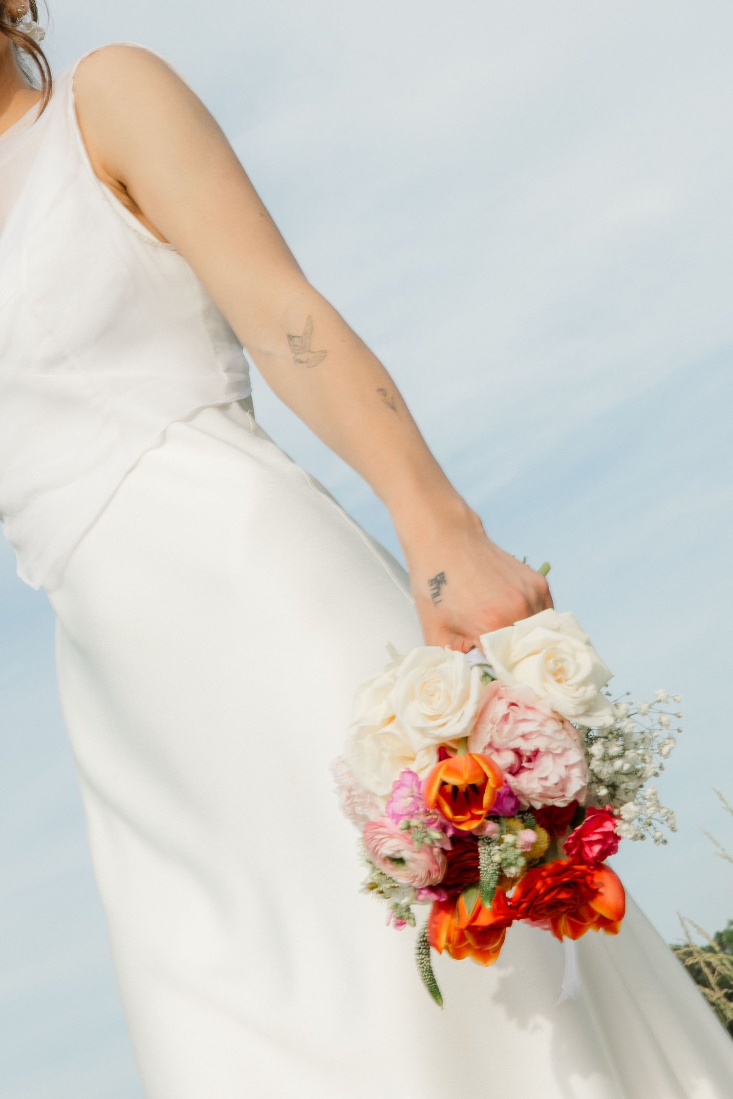 Bride in white gown holds vibrant bouquet of roses and ranunculus against a blue sky.