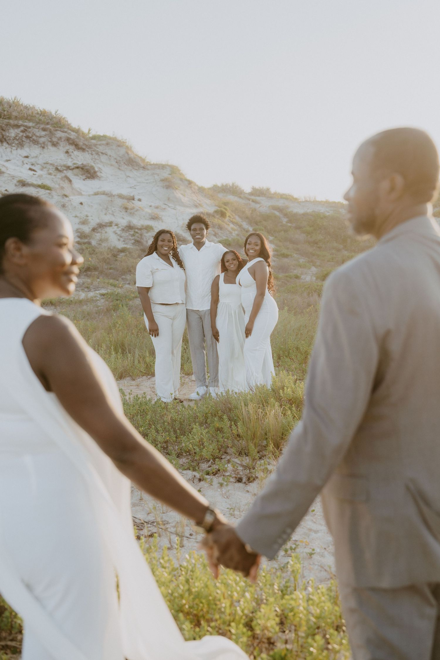 A couple holding hands on a sandy beach at sunset with wedding party members standing in background.
