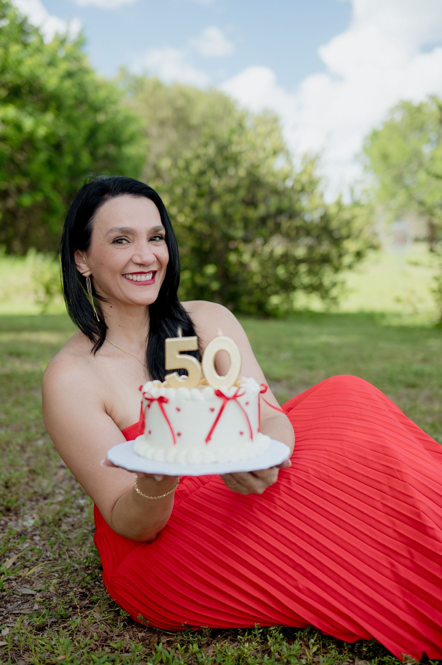 Woman in red flowing dress lying on grass holding a white birthday cake with gold number 50 decoration.
