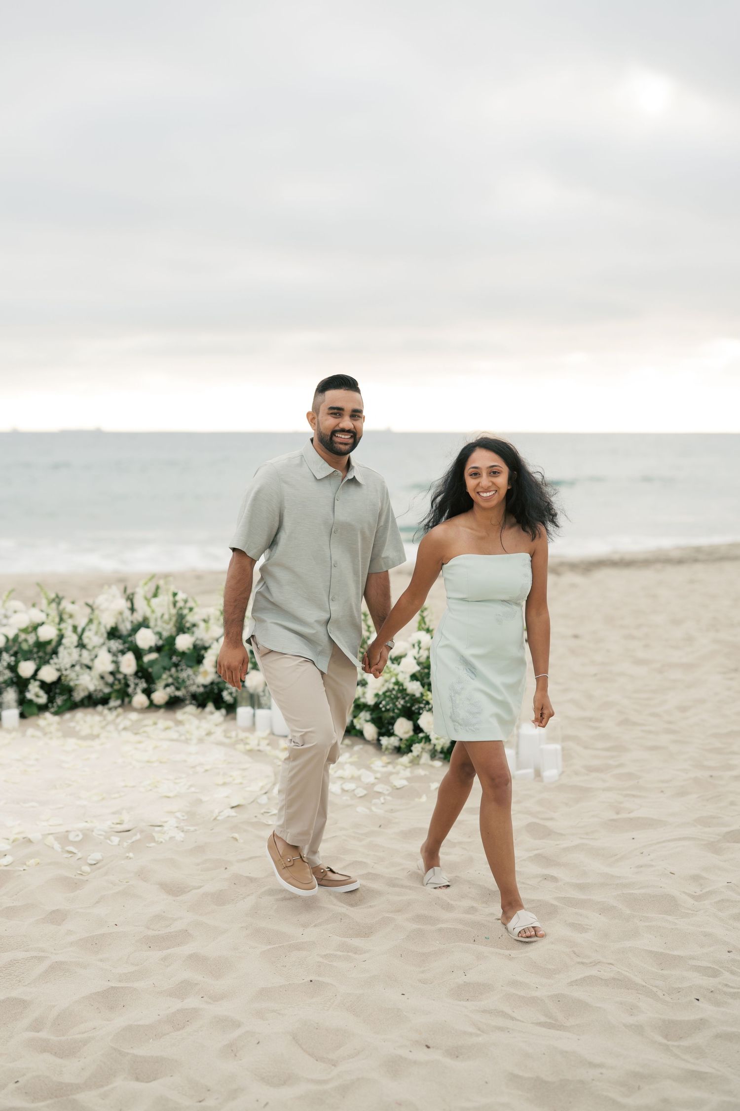A couple walks hand in hand on a sandy beach with white floral decorations and an overcast sky in the background.