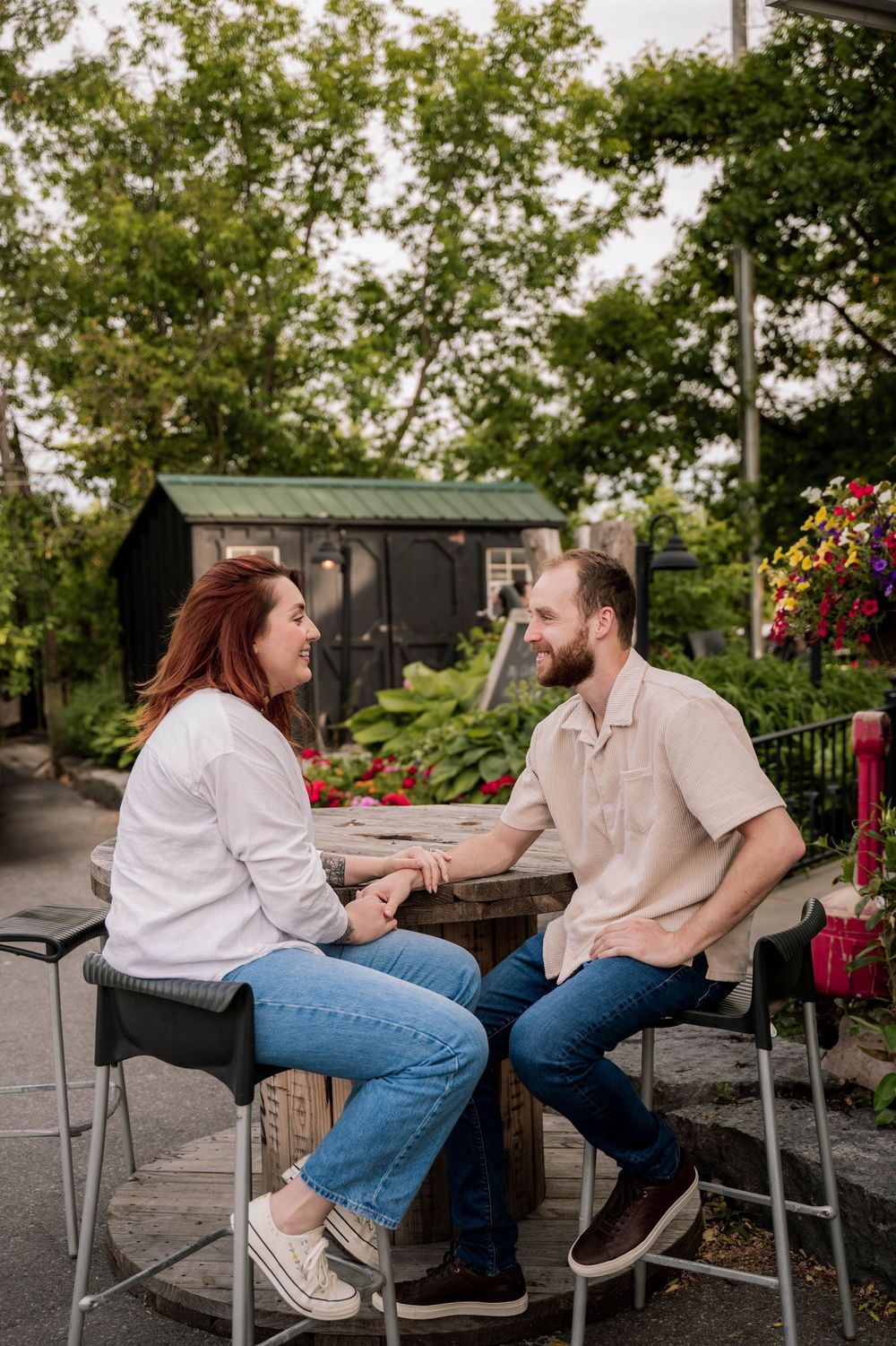 Two people sit on bar stools having an animated conversation at an outdoor patio with greenery and flowers in the background.