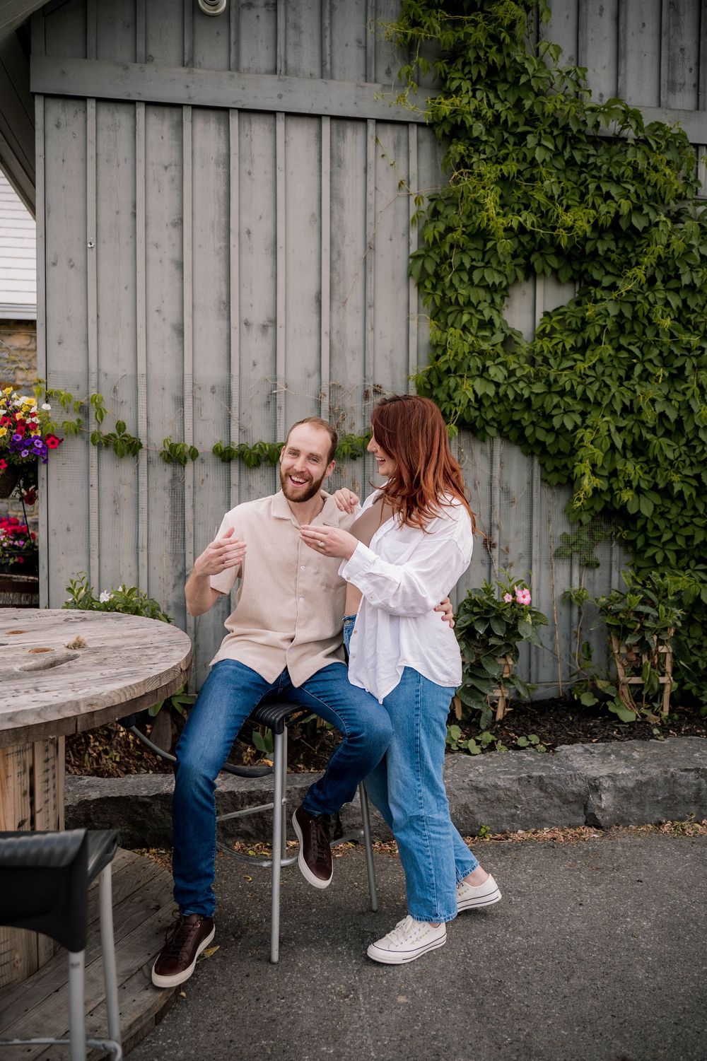 A couple sits together on a rustic bench outdoors against a grey concrete wall with climbing vines.