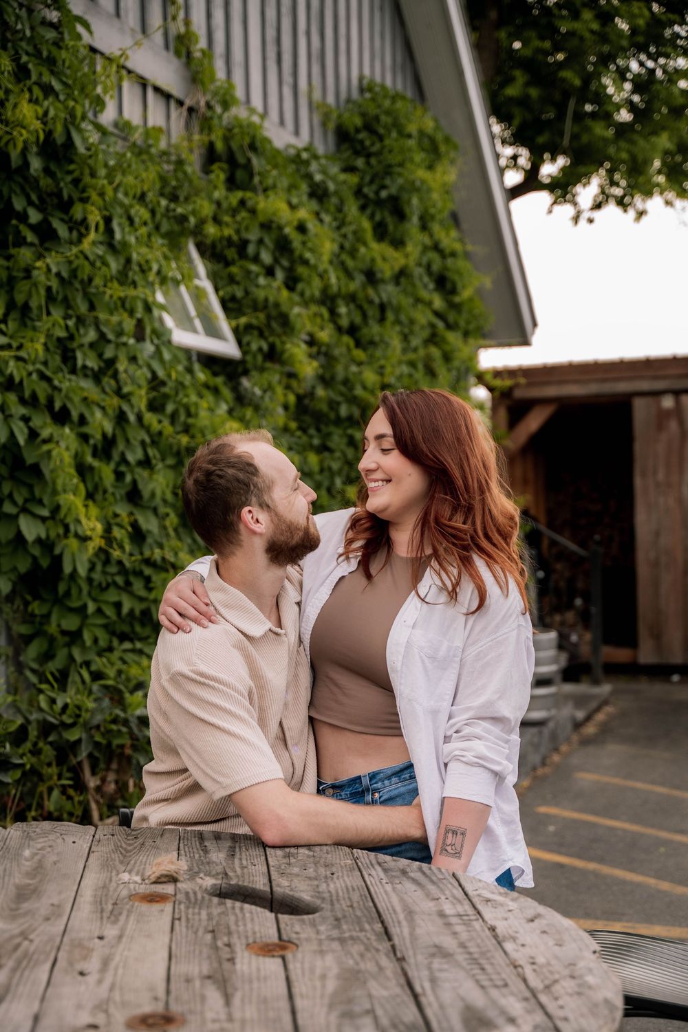 Couple sitting at rustic wooden table outside barn with ivy-covered wall in background sharing intimate moment.