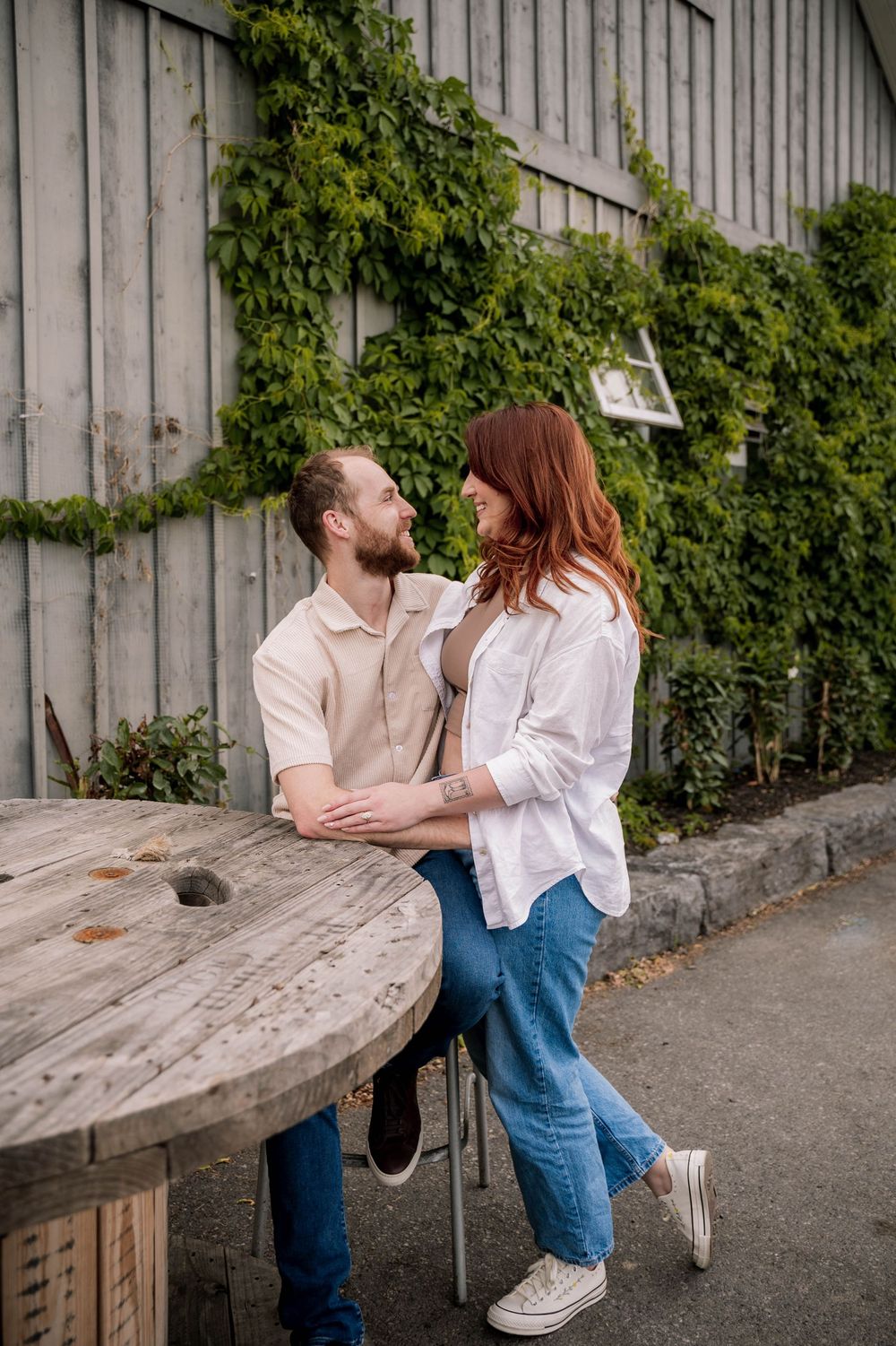 A couple shares a tender moment while standing by a rustic wooden table against an ivy-covered wall outdoors.