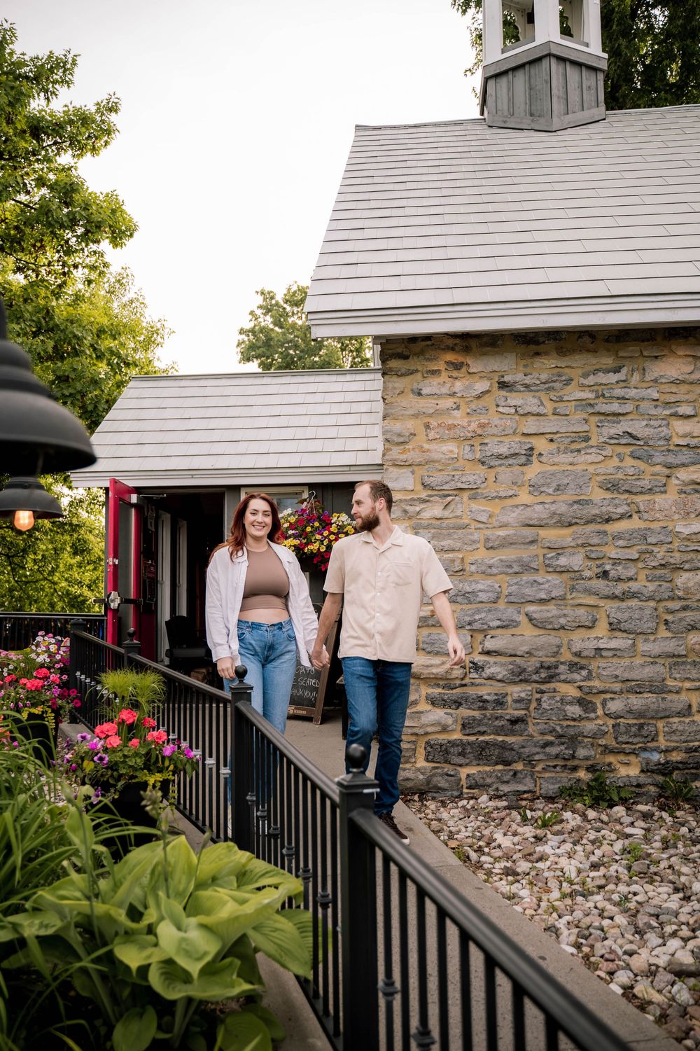 A couple walks together on a stone patio alongside a rustic stone building with flowers and landscaping.