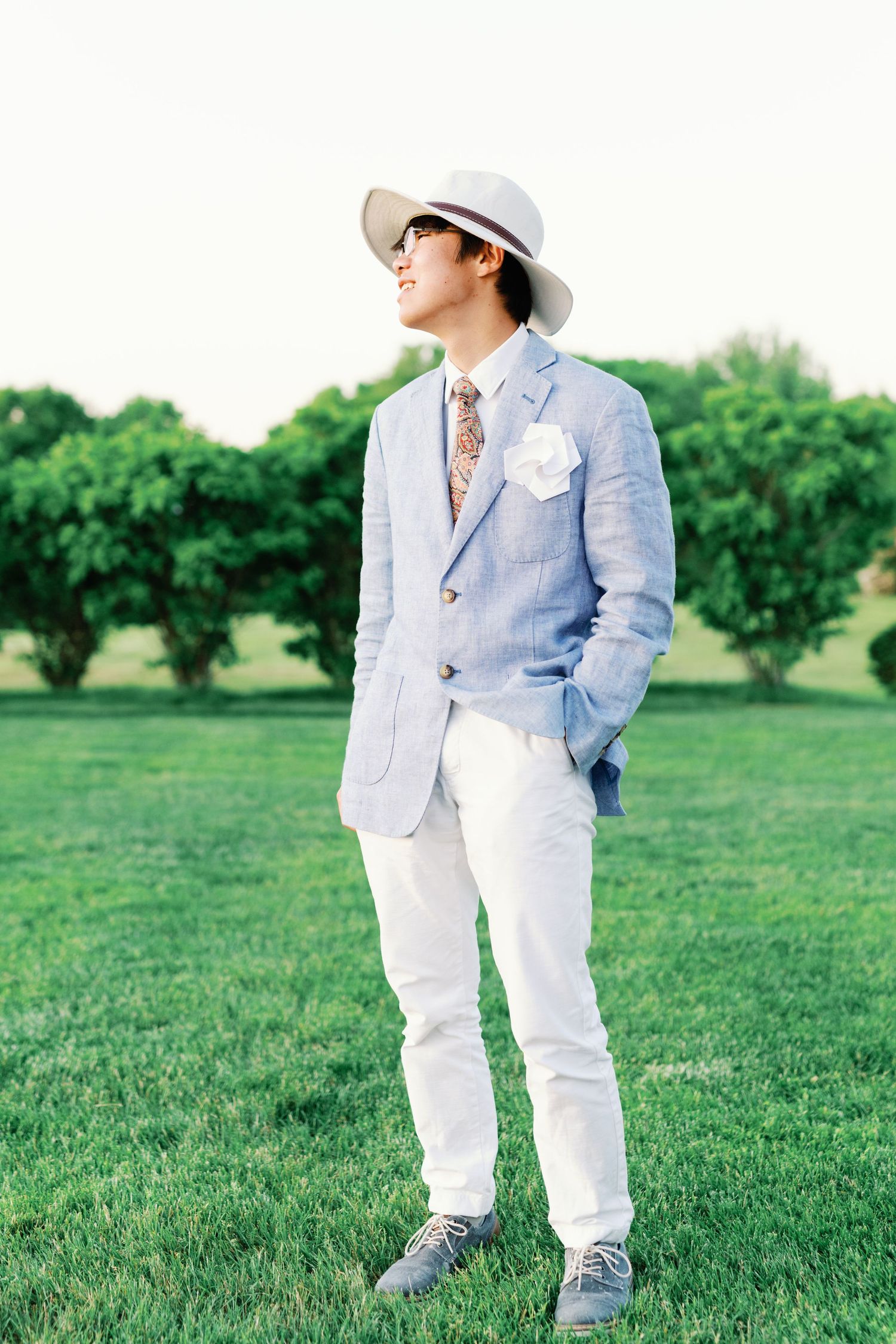 A well-dressed person in a light blue blazer, white pants, and panama hat stands on green grass in a garden setting.