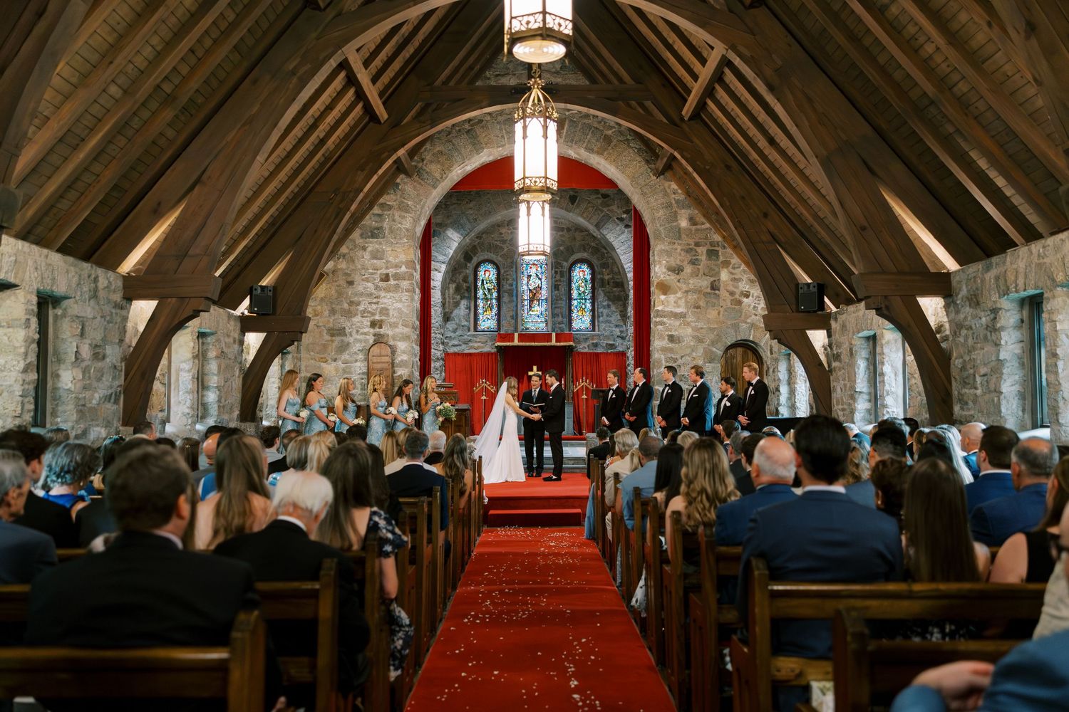A wedding ceremony takes place in a rustic stone church with vaulted wooden beams and a red carpet aisle.