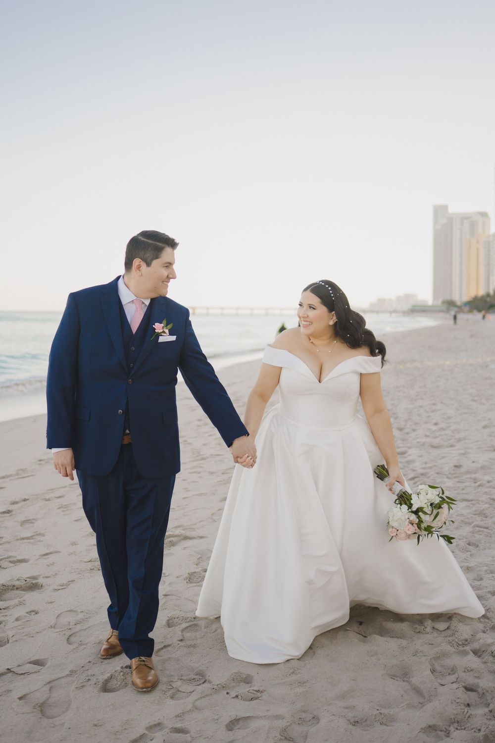 Newlywed couple walking hand in hand on a beach at sunset with a cityscape in the background.