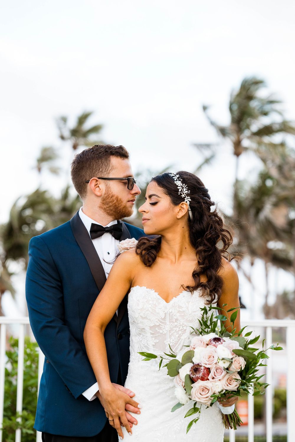 A romantic wedding couple shares an intimate kiss against a backdrop of palm trees on a sunny day.