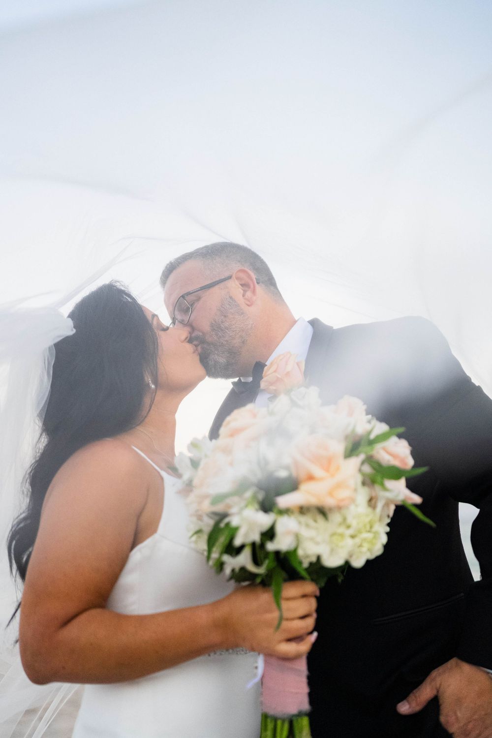 A romantic wedding photo of a couple sharing a kiss while holding a peach and white floral bouquet against bright sunlight.