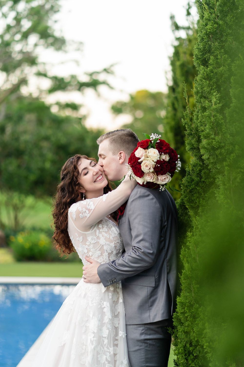 Romantic wedding couple embraces among green foliage beside blue water on their special day.
