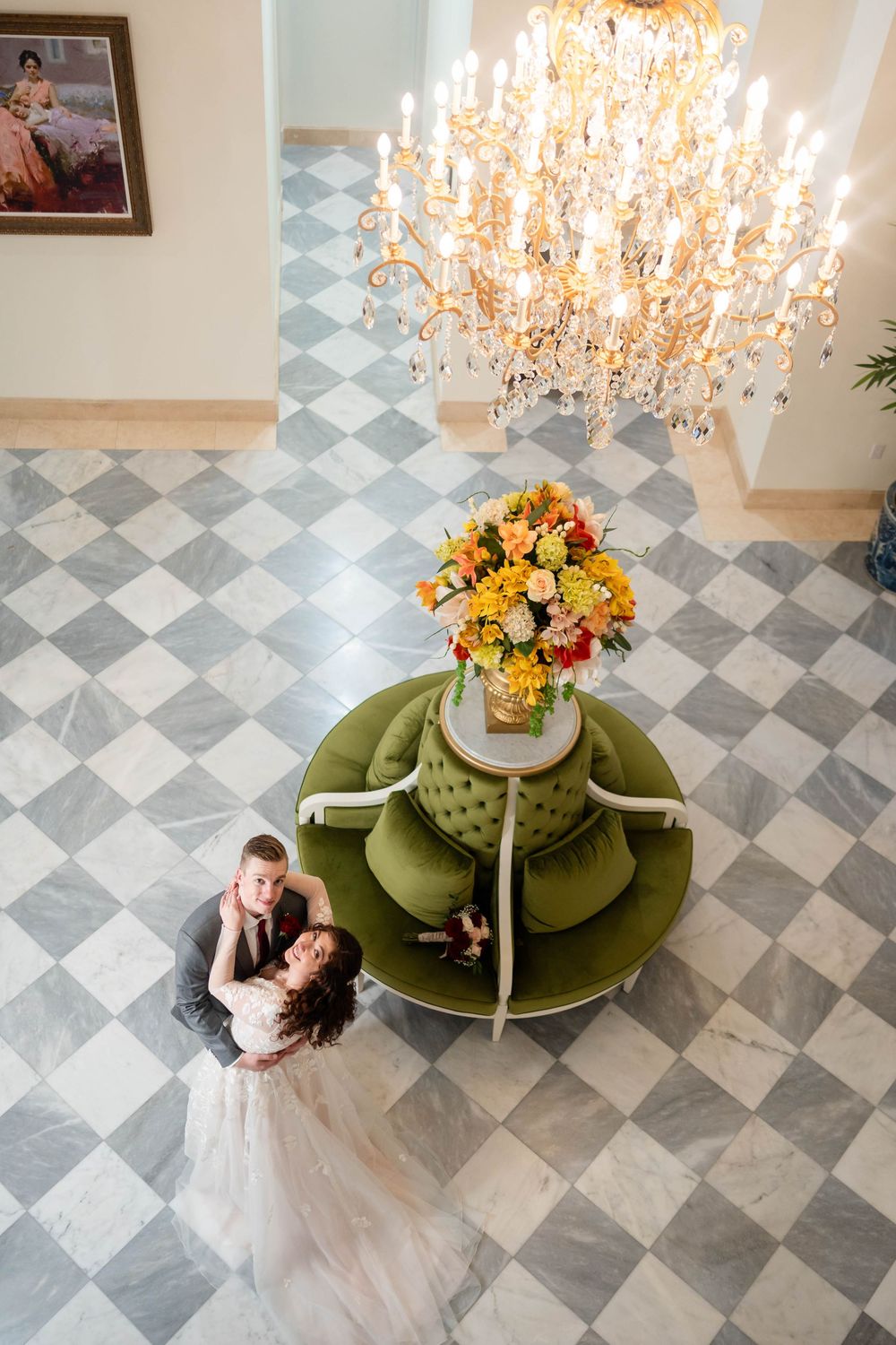 A bride and groom embrace on a checkered floor below an ornate crystal chandelier and orange floral arrangement.