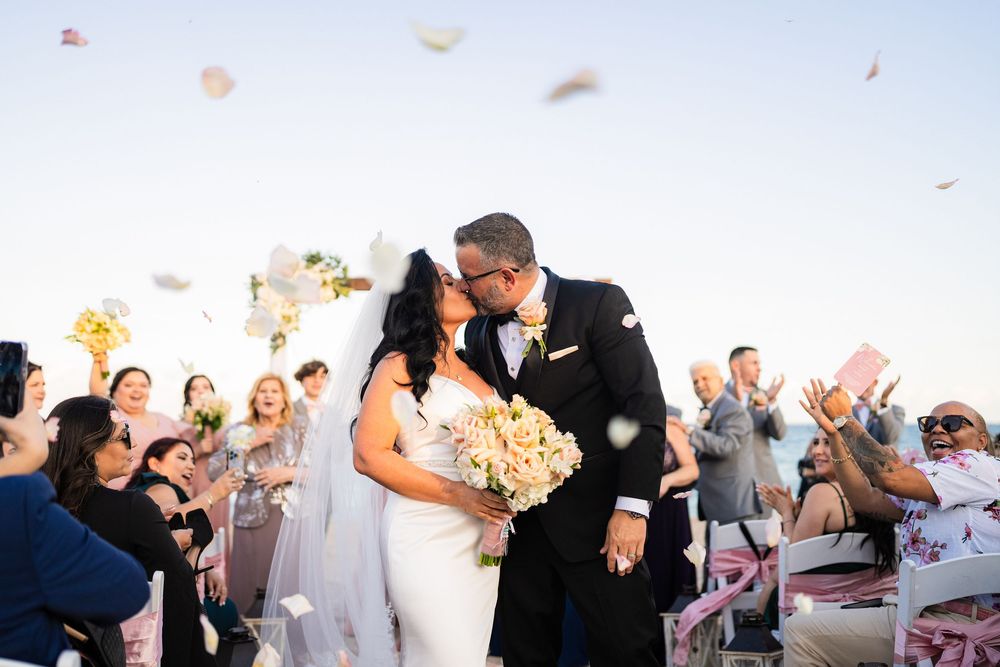 A romantic wedding celebration on the beach with flower petals floating in the air as guests cheer in the background.