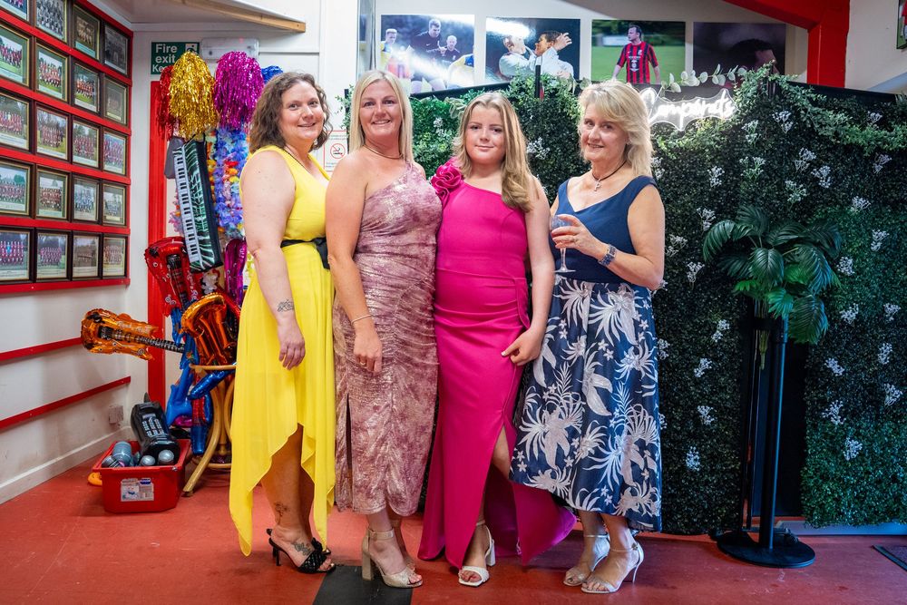 Four people in colorful formal dresses pose together at an event by a red wall with decorative foliage.