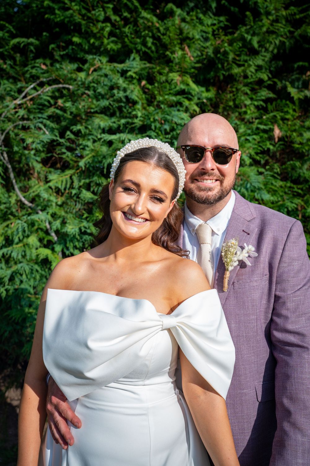 A couple poses outdoors on their wedding day against lush green foliage in summer light.