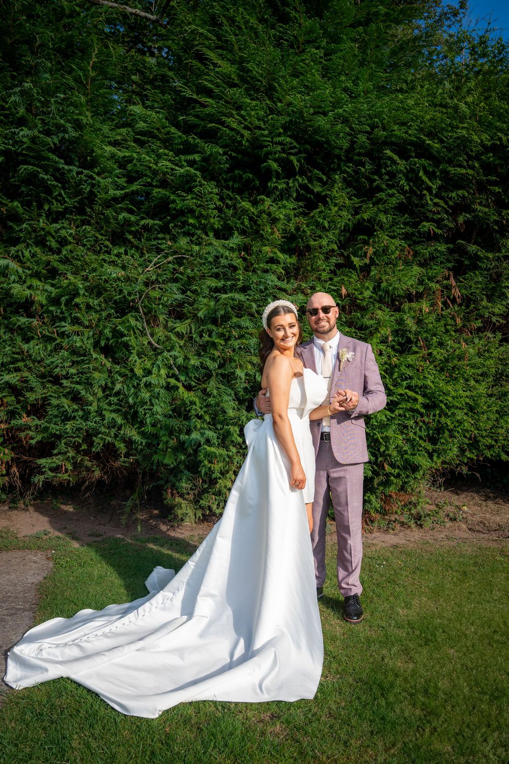 A couple poses together on green grass with lush foliage in the background as a white wedding dress train flows behind them.