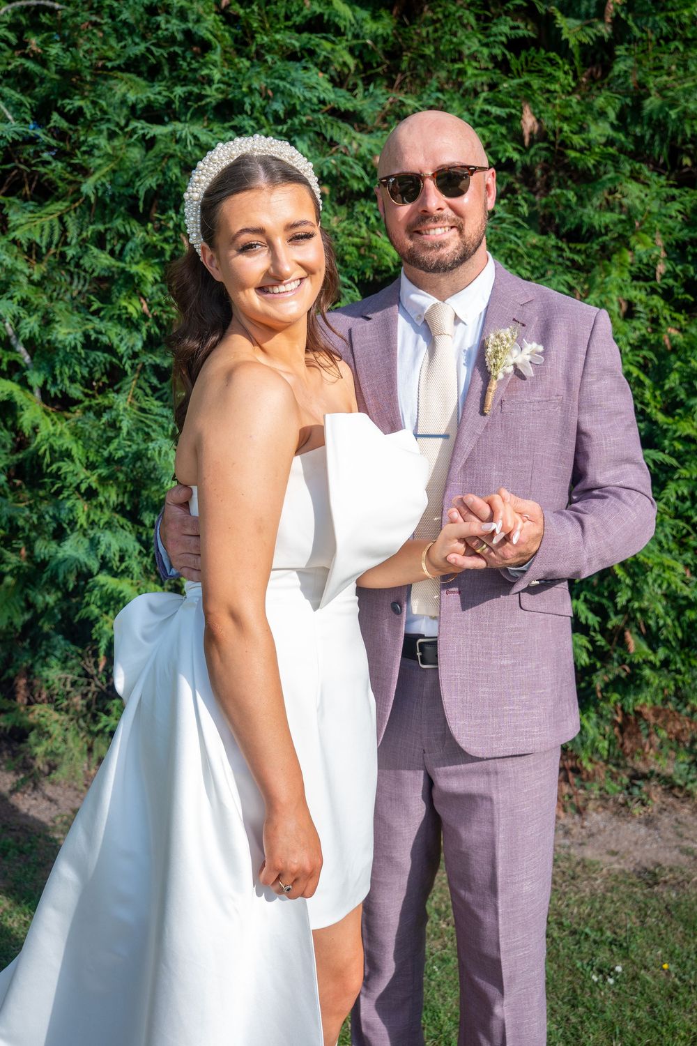 A wedding couple poses outdoors against green foliage, with the bride in white and groom wearing a dusty pink suit.
