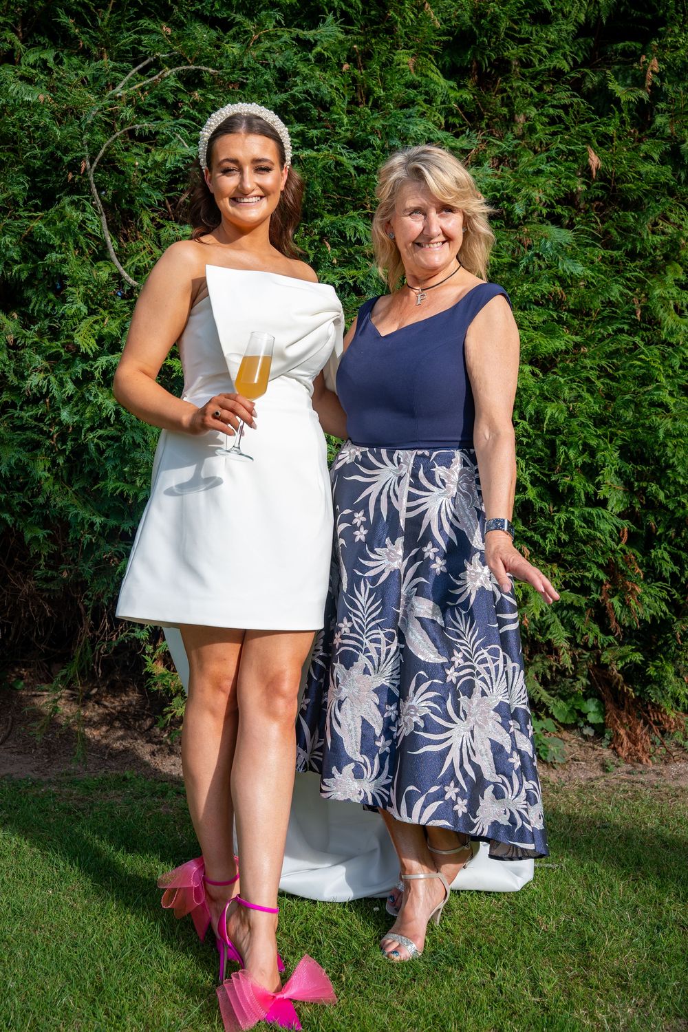 Two people in summer dresses pose together on a lawn, one wearing white and pink shoes, the other in navy and floral print.