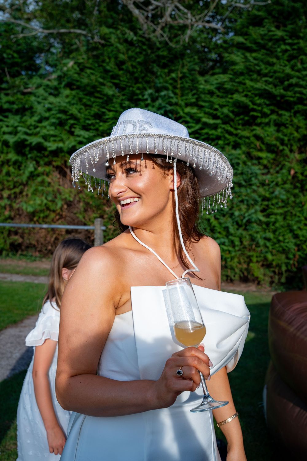 A joyful summer scene of someone in a white dress and sparkly fringe hat enjoying wine in a garden setting.