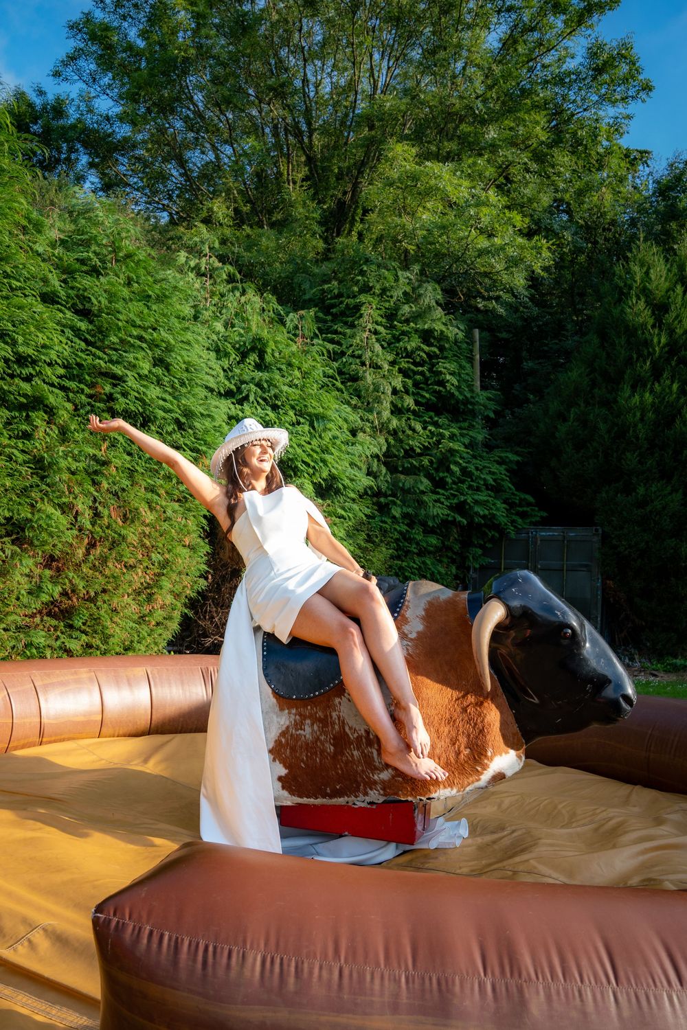 A person in a white dress rides a mechanical bull at an outdoor event on a sunny summer day.