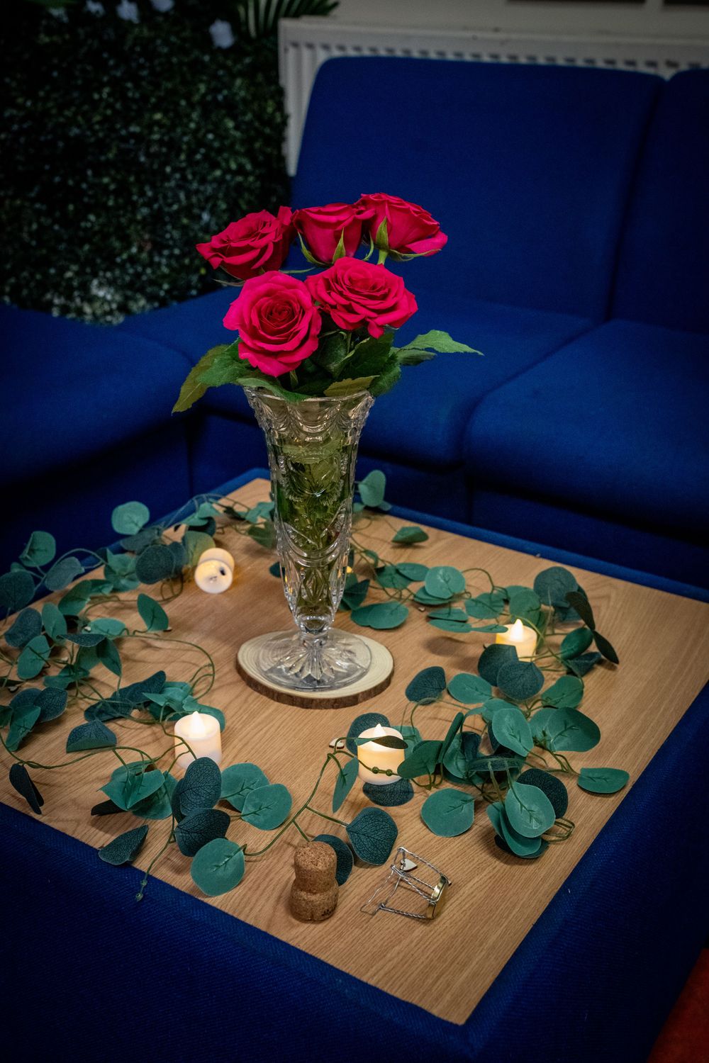 Red roses in a crystal vase with eucalyptus leaves and candlelight on a wooden table against navy blue decor.