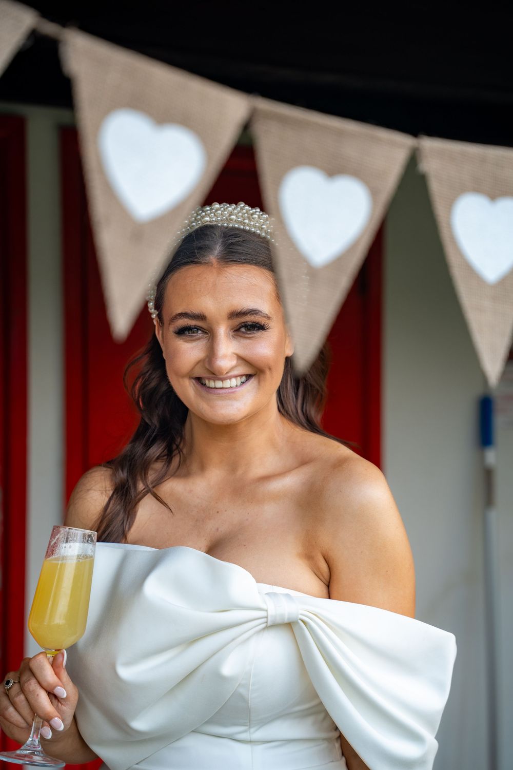 A bride in a white off-shoulder dress with a tiara holds a mimosa while smiling in front of heart-shaped bunting.