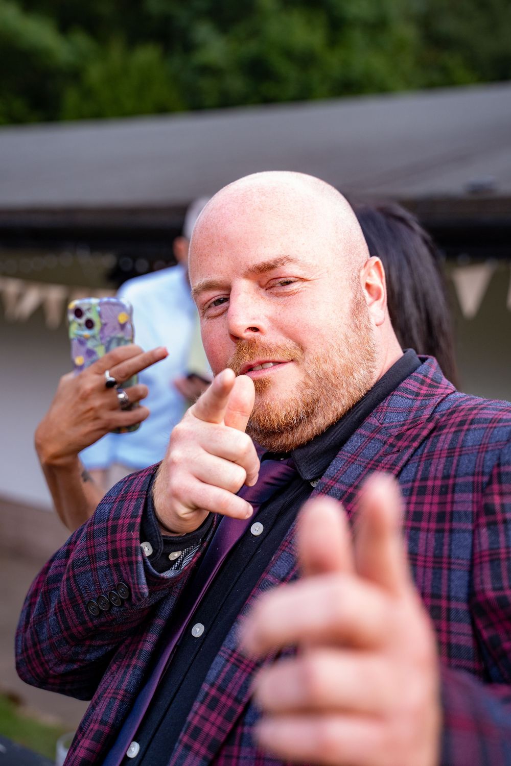 Person in plaid shirt pointing at camera while someone holds up phone in background during outdoor gathering.