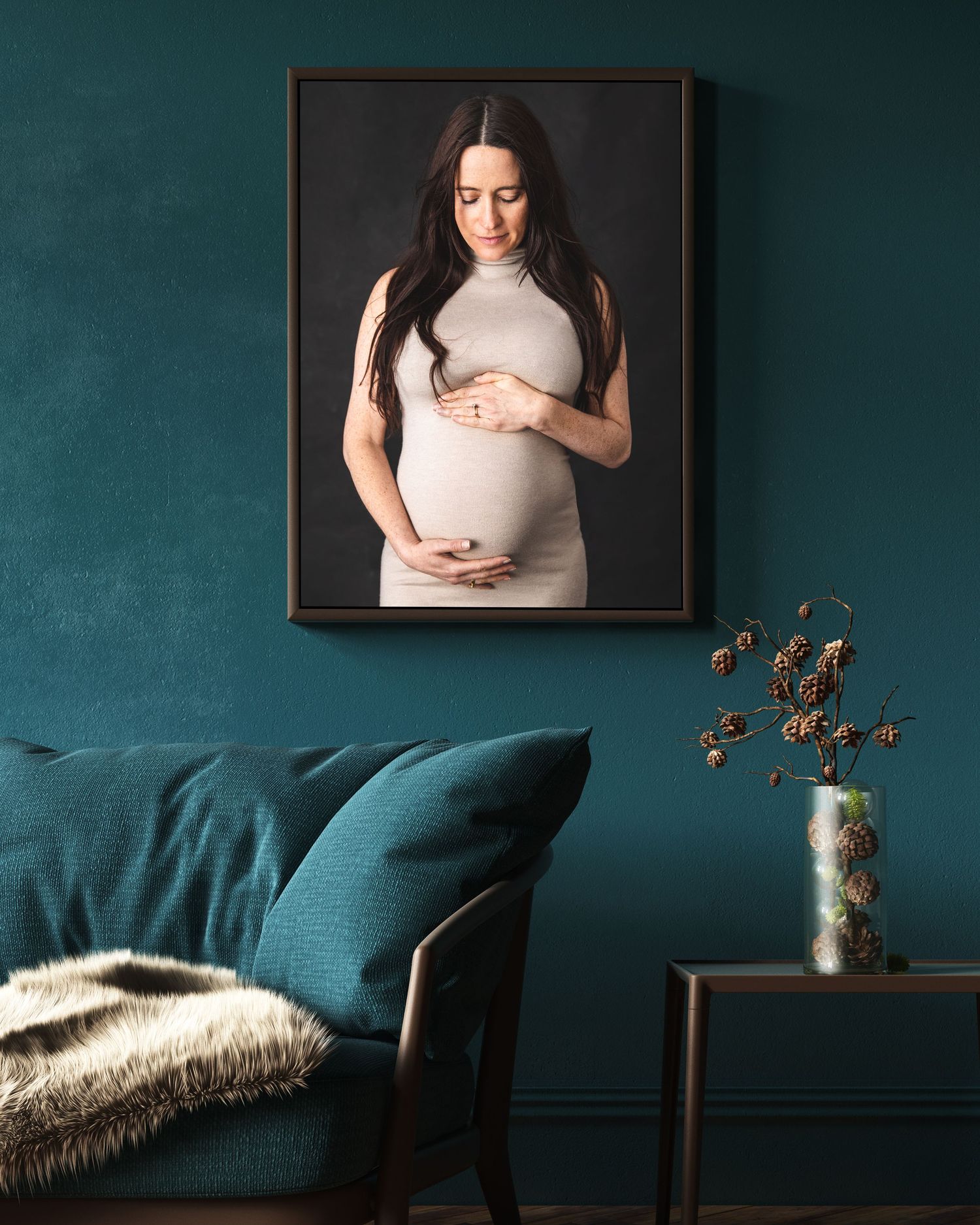Maternity portrait displayed on teal wall above cozy velvet sofa with fur throw and decorative vase on side table.