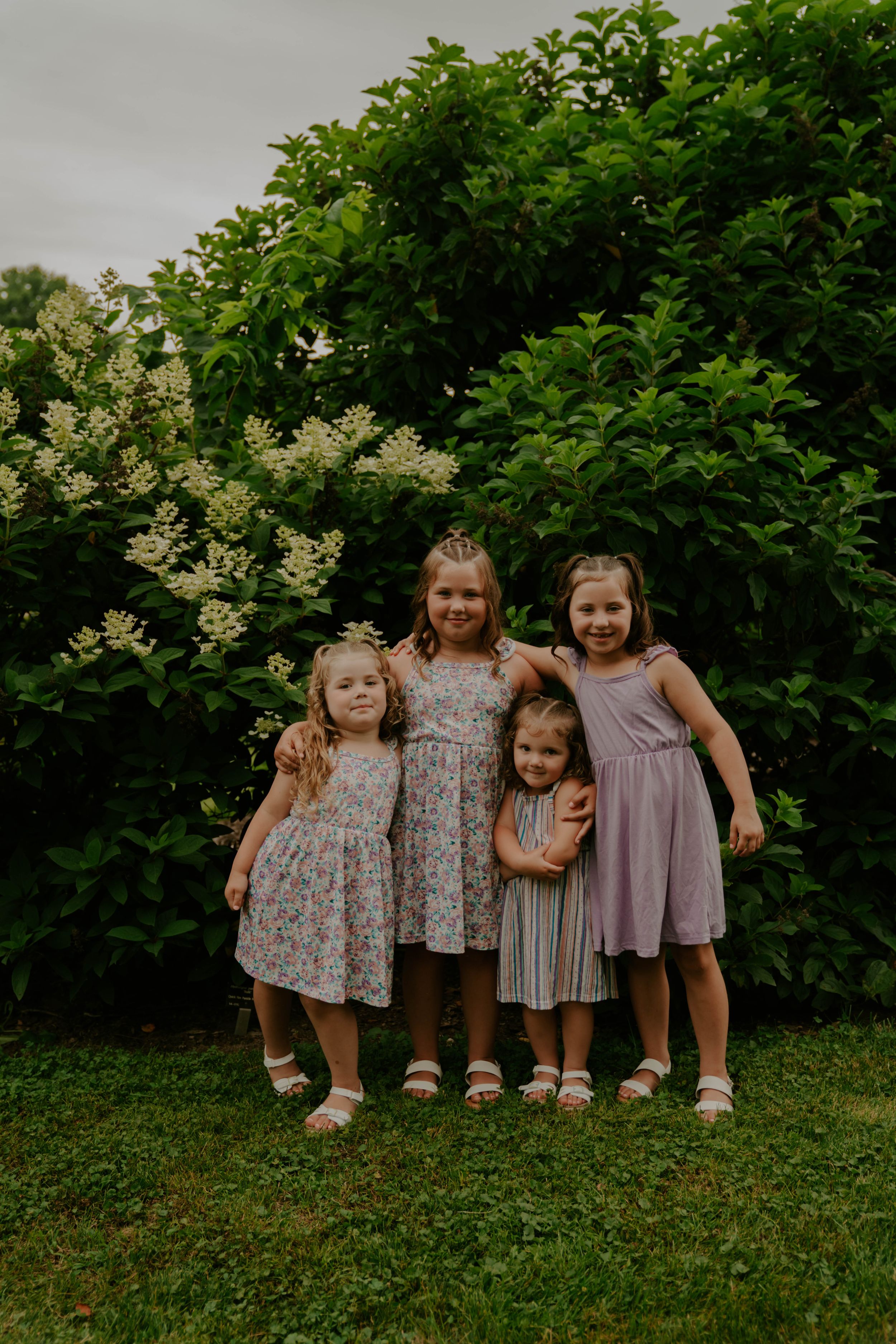 Children in matching summer dresses posing together on lawn near flowering bushes.