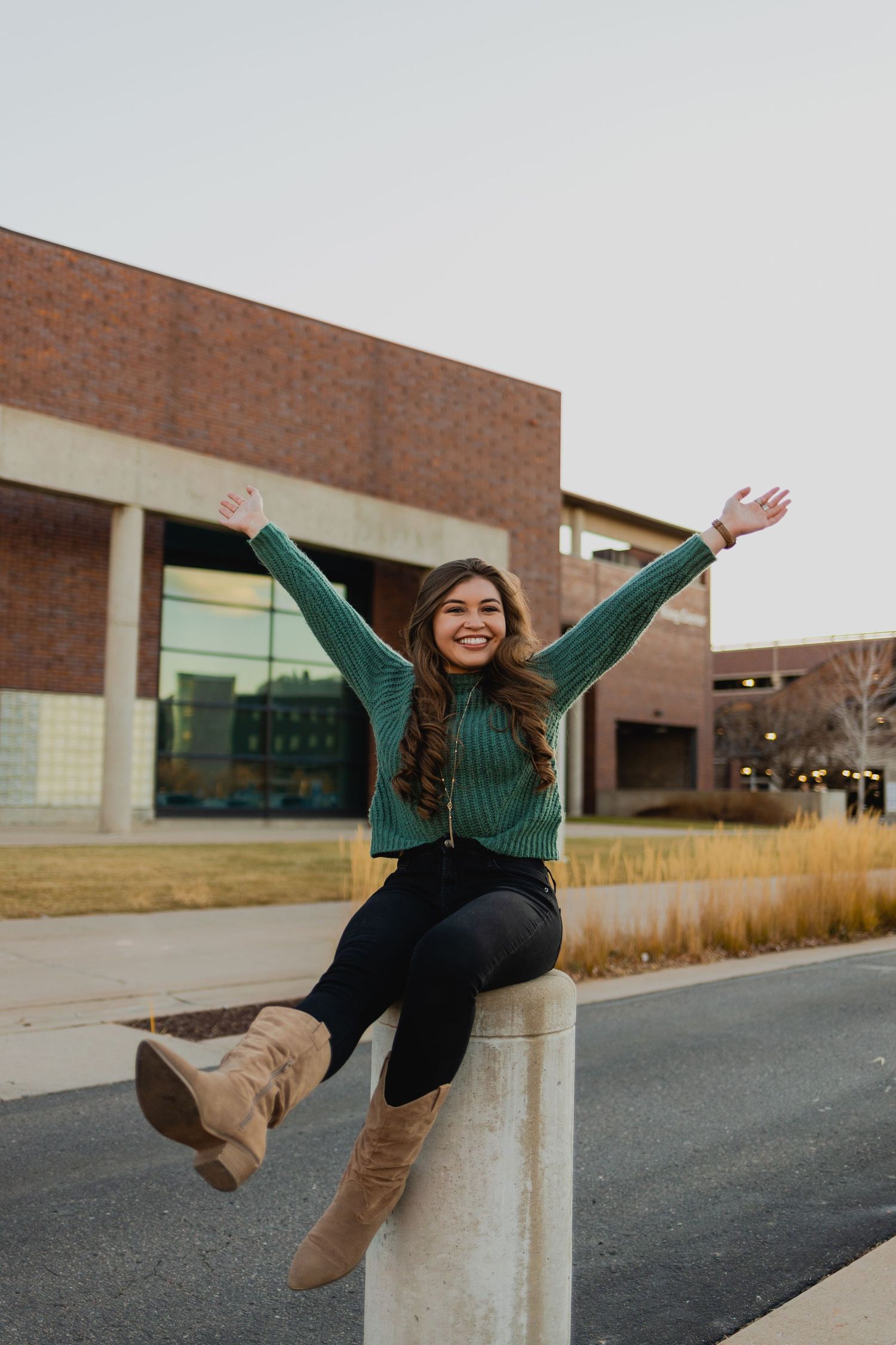 Person in green sweater and black pants does playful jumps and poses against a brick building exterior.