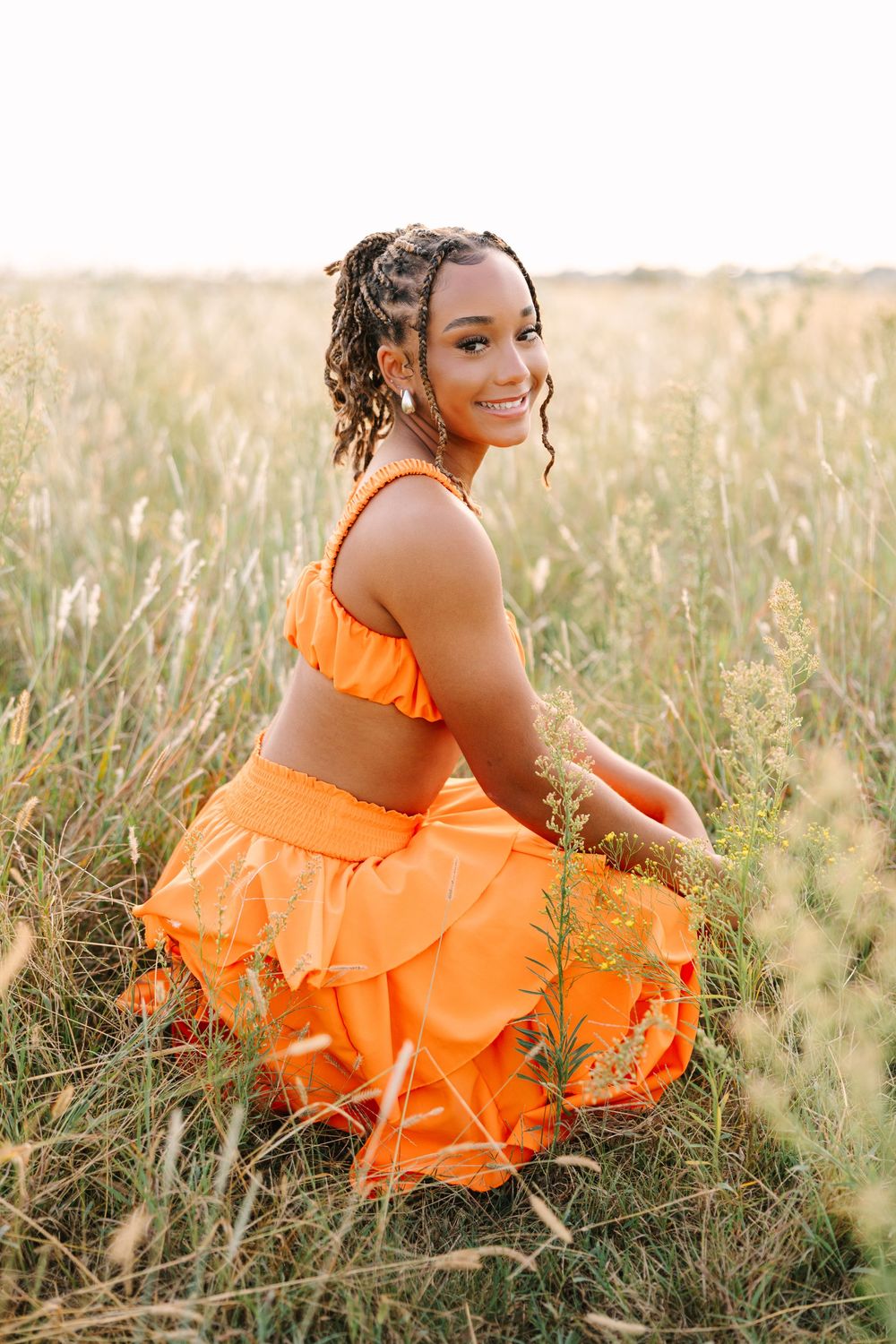 A person in a bright orange ruffled dress poses gracefully in a sunlit field of tall grass during golden hour.