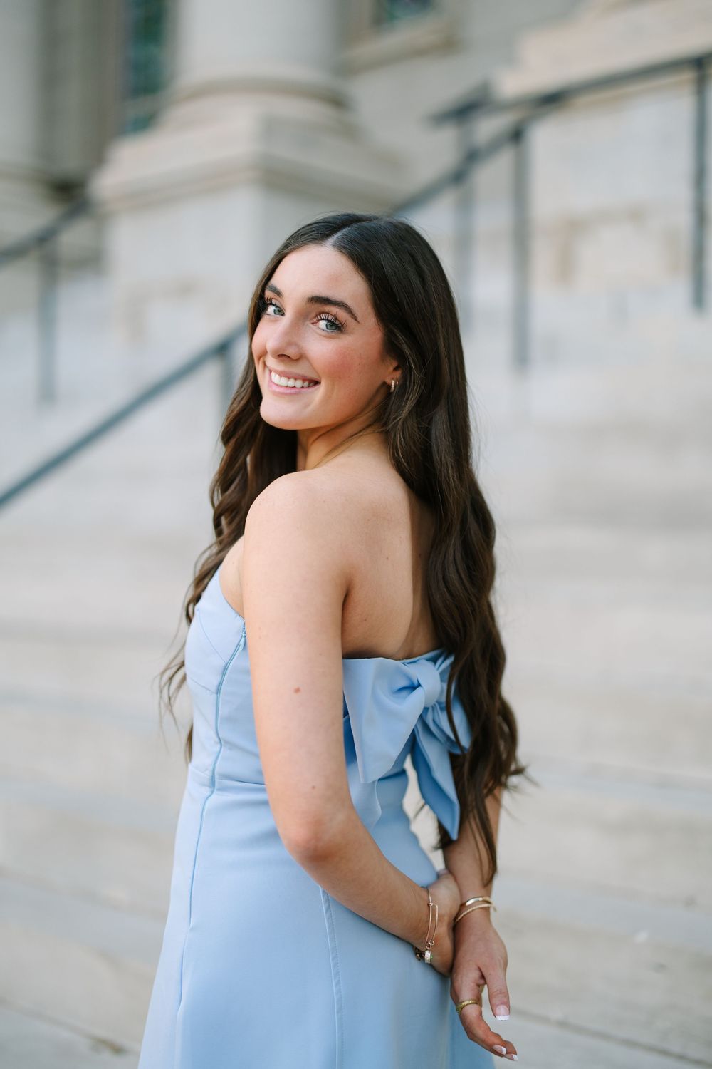Young woman in a strapless light blue gown with bow detail, smiling over her shoulder on elegant stone steps.
