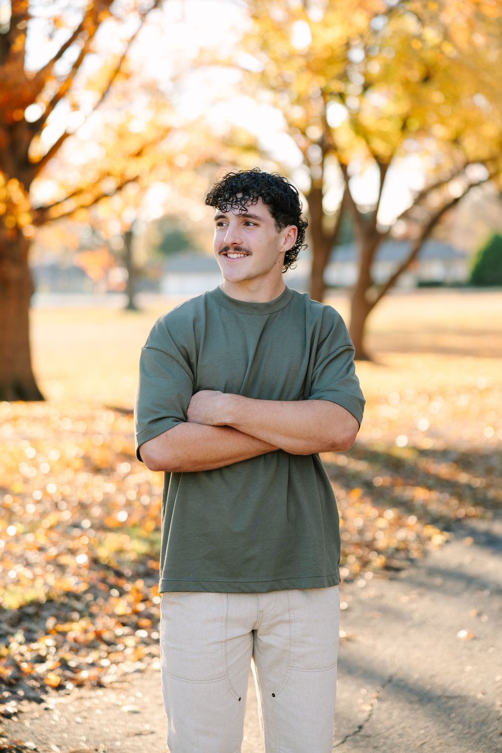 Person wearing a green t-shirt and light pants stands in a park with golden autumn trees in the background.